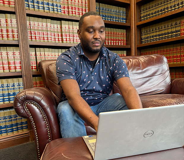 A student sitting at a desk with a laptop open in front of bookshelves.